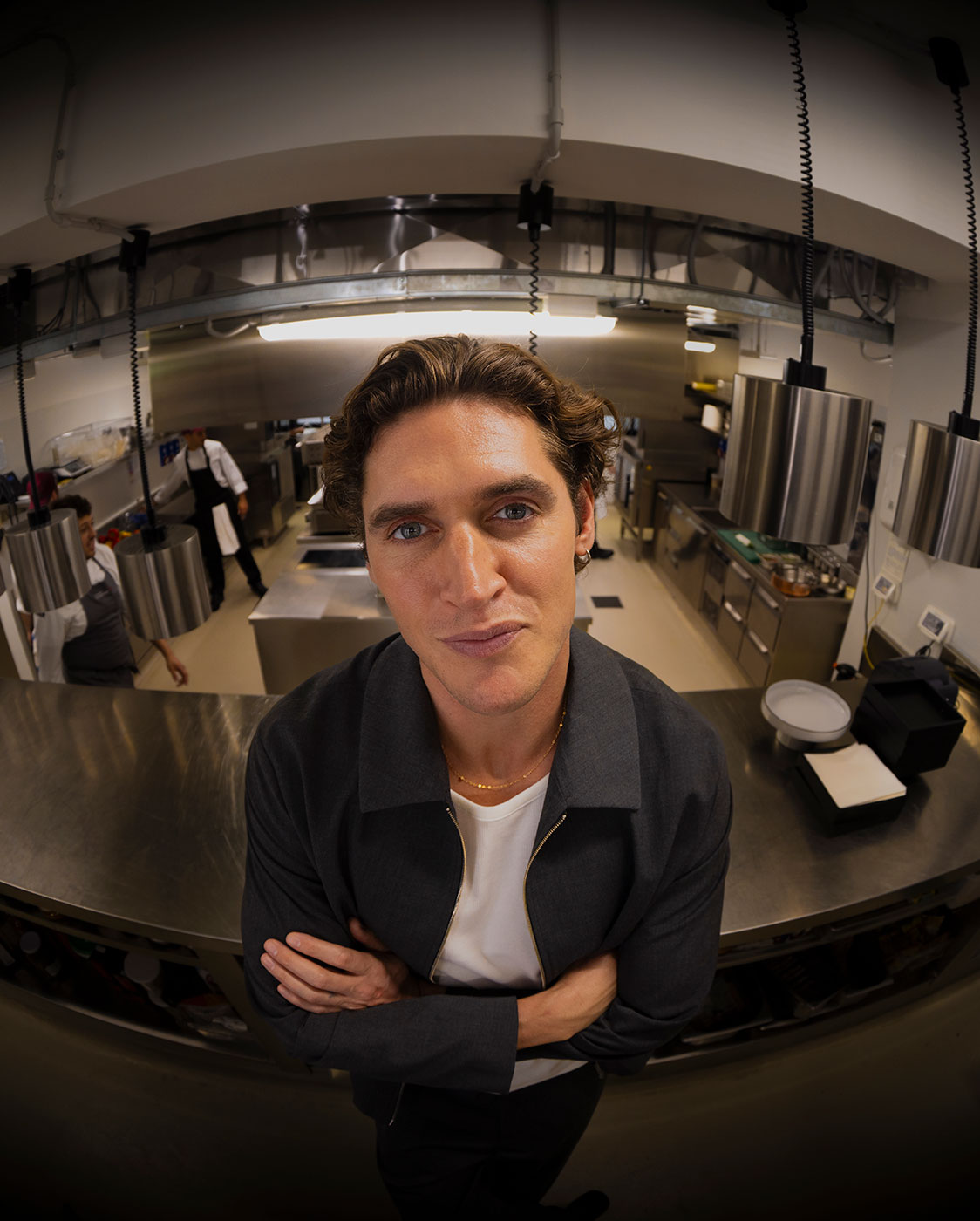 Ein Mann mit gewellten braunen Haaren und blauen Augen steht in einer modernen Industrieküche und blickt mit verschränkten Armen und einem leichten Lächeln in die Kamera. Er trägt eine dunkle Jacke über einem weißen Hemd. Um ihn herum stehen Geräte aus rostfreiem Stahl.