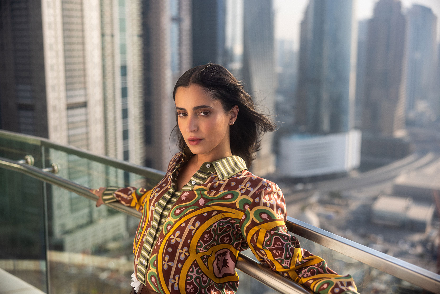 Una mujer de pelo largo y oscuro, con una camisa estampada de colores, se apoya en la barandilla de un balcón acristalado con el horizonte de una ciudad y altos edificios difuminados al fondo.