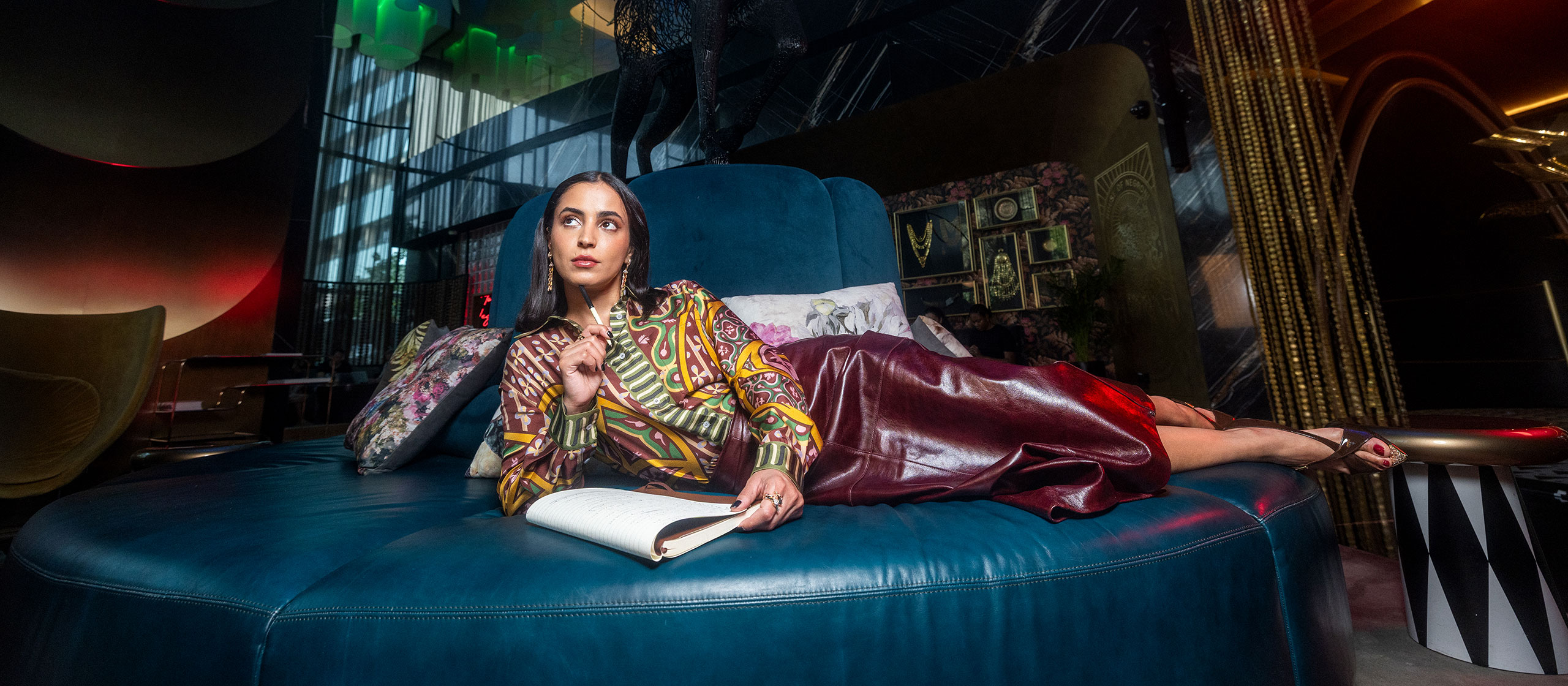 Eine Frau in bunter Bluse und kastanienbraunem Rock liegt auf einer großen runden blauen Couch, hält einen Stift und ein Notizbuch in der Hand und schaut nachdenklich zur Seite in einem stilvollen, modernen, schwach beleuchteten Raum mit eklektischem Dekor.