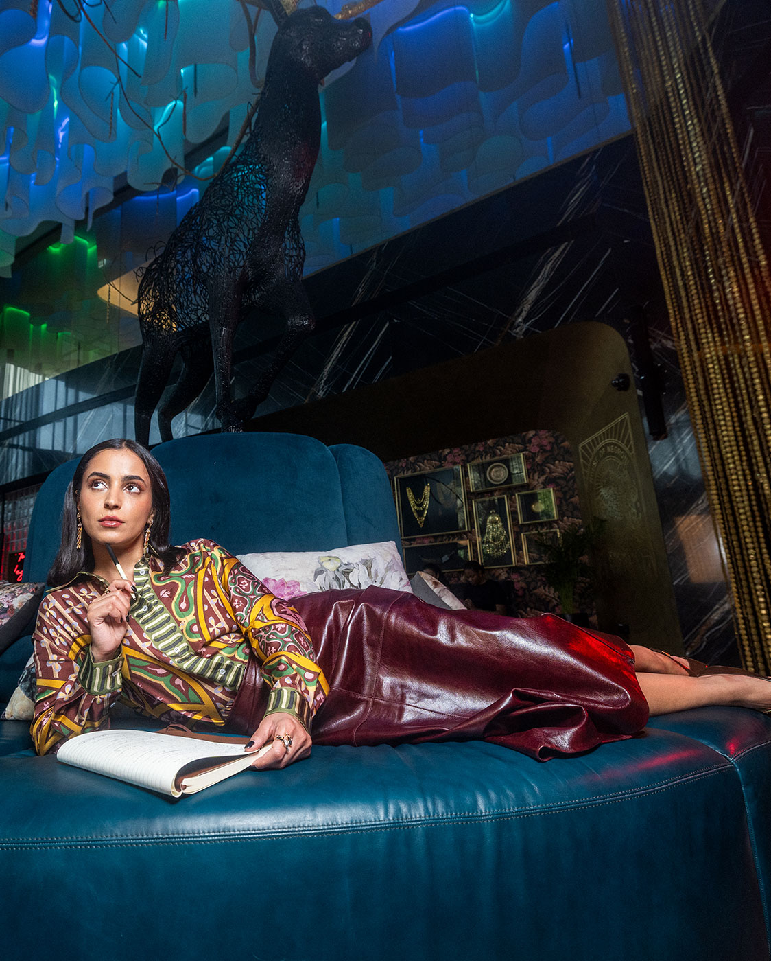 Eine Frau in bunter Bluse und langem rotem Rock sitzt auf einem großen blauen Stuhl, in der Hand einen Bleistift und ein aufgeschlagenes Notizbuch. In dem modernen, stilvollen Interieur sind dekorative Leuchten und eine schwarze Hirschstatue zu sehen.