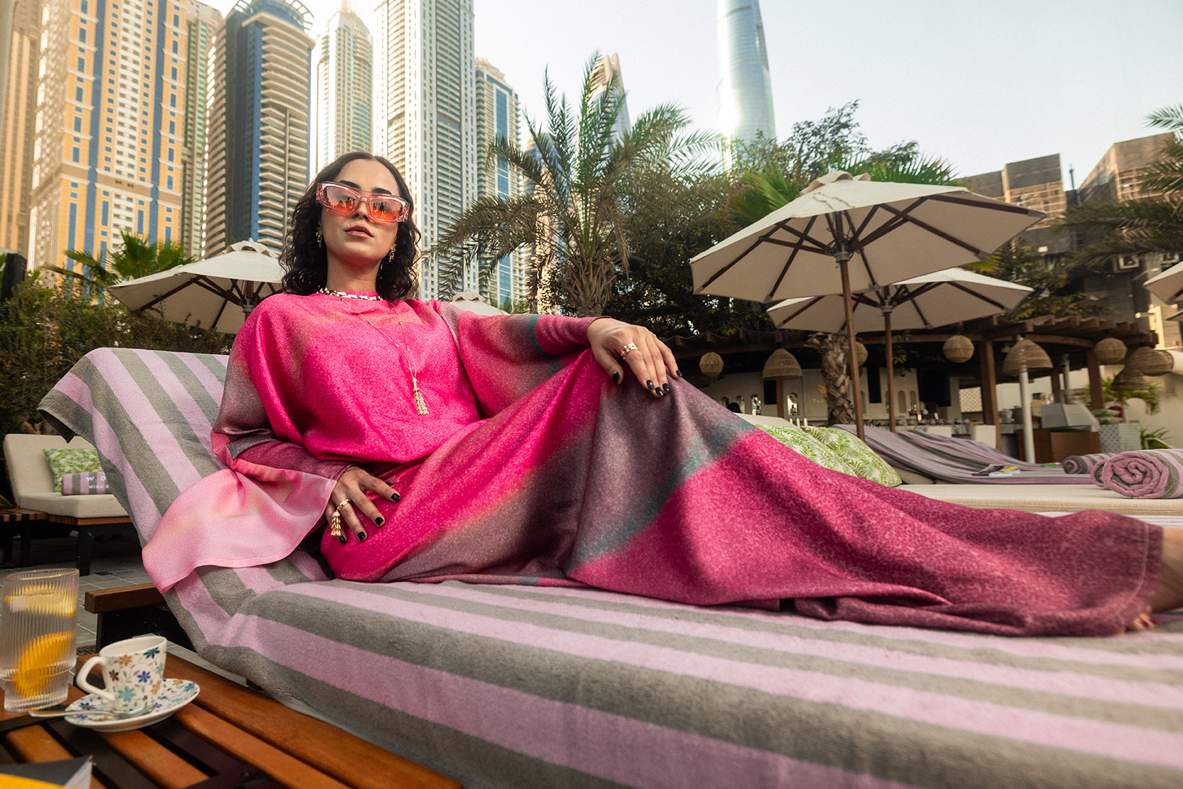 Una mujer vestida con un elegante traje rosa y morado descansa en una silla a rayas junto a la piscina, con gafas de sol y joyas, con rascacielos, palmeras y sombrillas de fondo. En una mesa cercana hay una copa y un vaso.