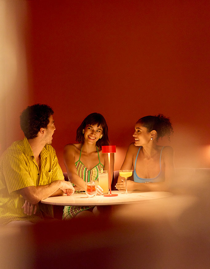 Drei Personen sitzen um einen kleinen runden Tisch mit Getränken, lächeln und unterhalten sich in einem warm beleuchteten Raum mit orangefarbenen Wänden und einer roten Lampe in der Mitte.