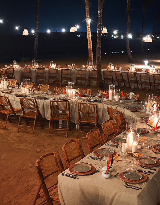 Abendessen im Freien an einem nächtlichen Sandstrand mit langen, mit beigen Tischdecken bedeckten Tischen, Holzstühlen, Tellern, Gläsern, Kerzen und Lichterketten, die eine warme, festliche Atmosphäre schaffen.