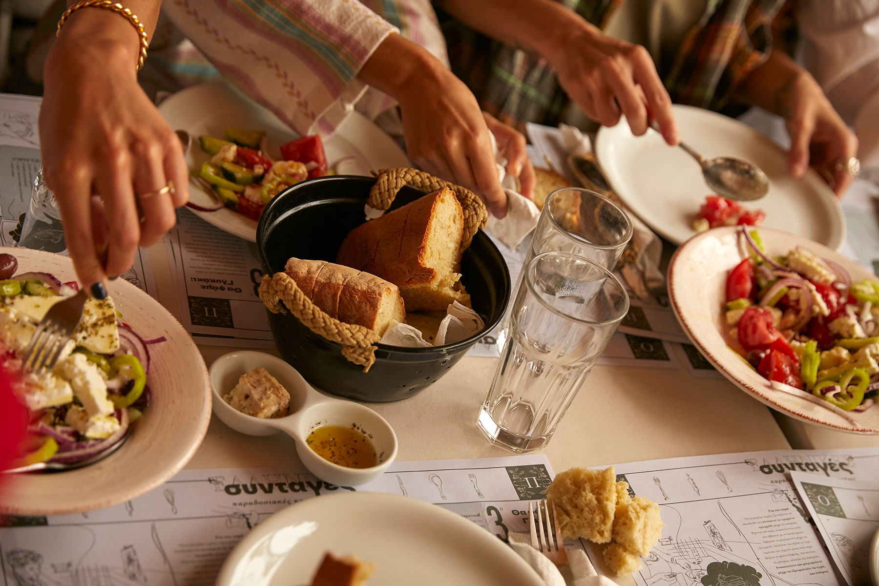 Nahaufnahme von mehreren Personen, die an einem Tisch mit Tellern mit griechischem Salat, Brot in einem Korb, Dips und Wassergläsern essen, wobei die Hände nach dem Essen greifen.