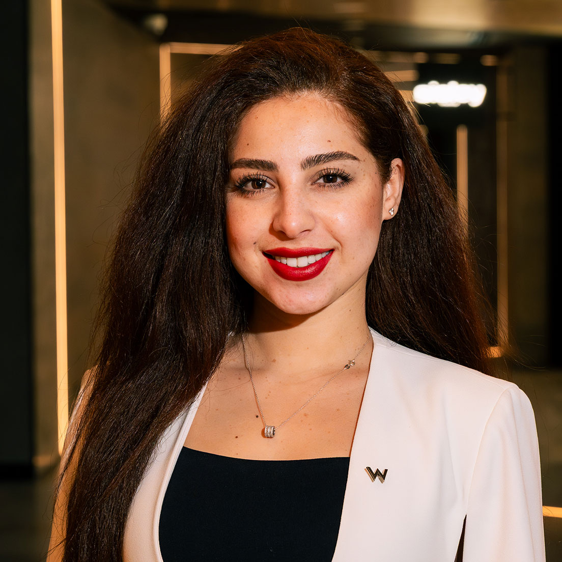 Una mujer con el pelo largo y castaño y los labios pintados de rojo sonríe a la cámara. Lleva una americana blanca sobre un top negro y un collar, en un interior moderno y cálidamente iluminado.