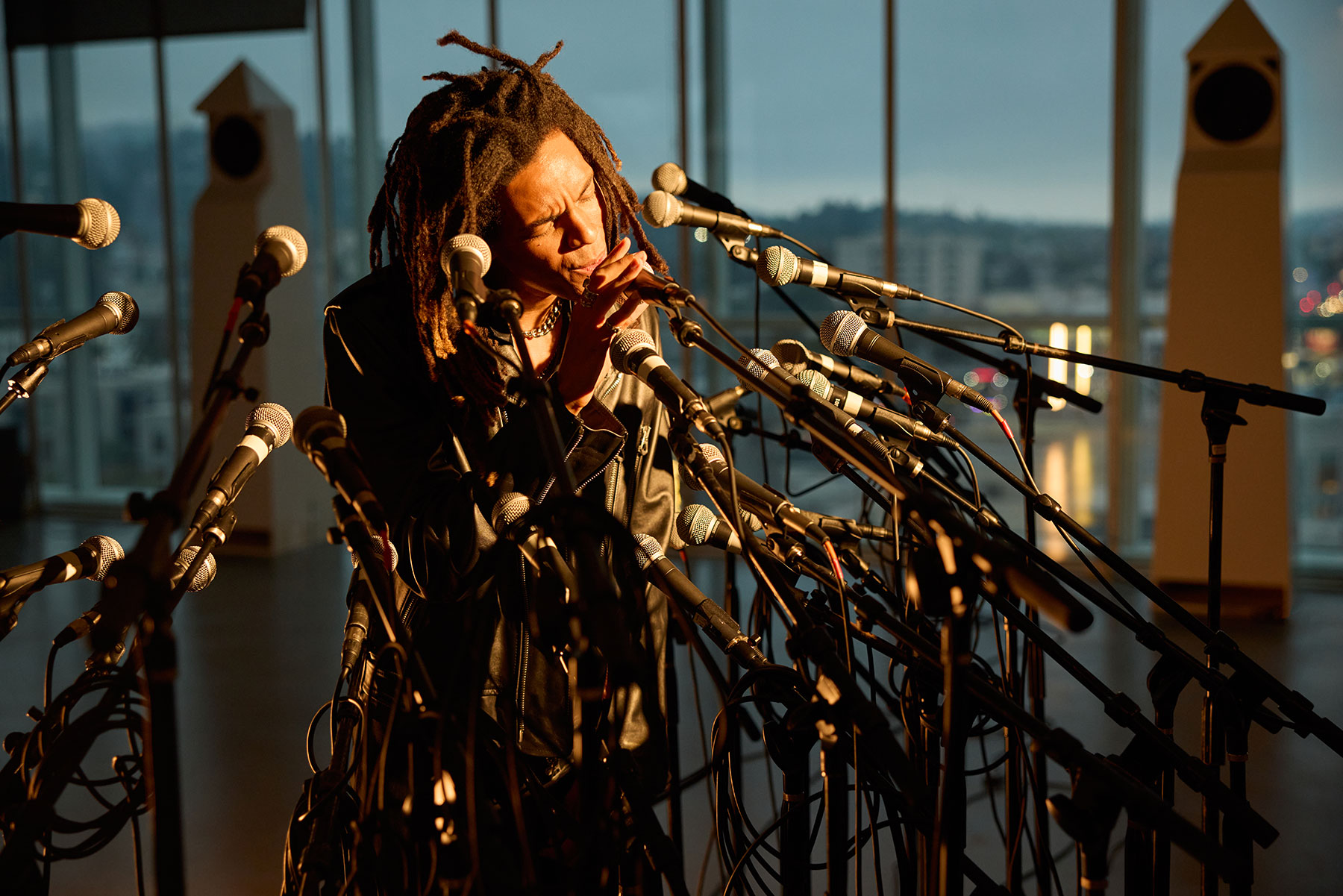 Eine Person mit Dreadlocks spielt vor vielen Mikrofonen Mundharmonika in einem sonnendurchfluteten Raum mit großen Fenstern und Blick auf eine Stadtlandschaft.