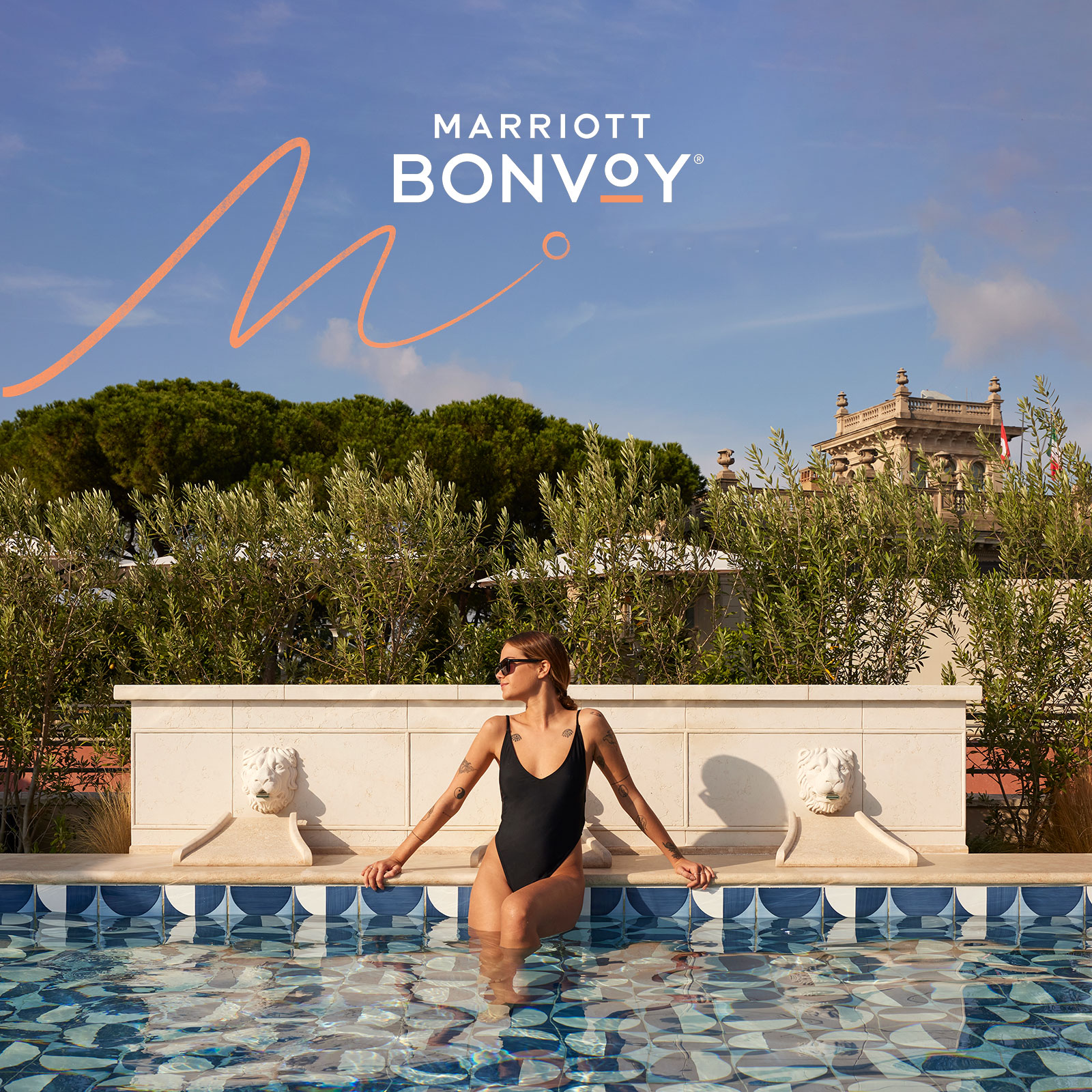 Eine Frau in einem schwarzen Badeanzug sitzt am Rande eines gekachelten Außenpools mit Löwenkopfbrunnen, umgeben von Grün. Über ihr ist unter blauem Himmel das Marriott-Bonvoy-Logo zu sehen.