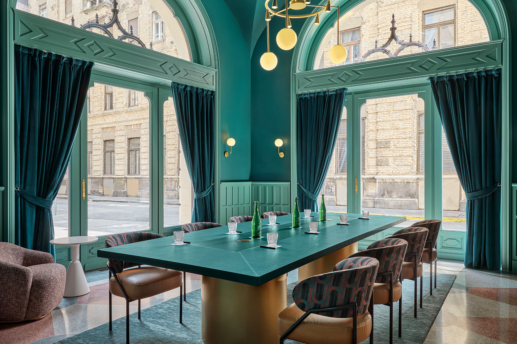 Ein stilvoller, moderner Essbereich mit tealfarbenen Wänden und raumhohen Fenstern, gewölbten Türen, eleganter Beleuchtung und einem großen grünen Tisch mit gemusterten Stühlen. Blaue Vorhänge rahmen die Fenster ein und geben den Blick auf ein Backsteingebäude frei.
