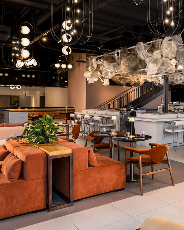 Ein modernes Restaurant mit rostfarbenen Sofas, Holzstühlen, einer Marmortheke mit hohen Hockern, eleganten Beleuchtungskörpern und skulpturalen wolkenartigen Deckendekorationen. Die Tische sind mit Tellern und Gläsern gedeckt.