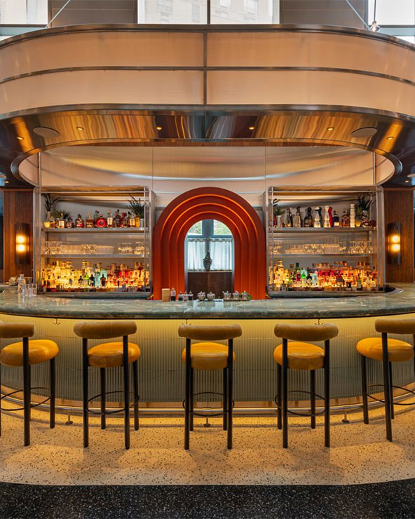 Eine moderne Bar mit einem geschwungenen Tresen, gelben, gepolsterten Hockern und dahinter liegenden Flaschenregalen. Warme Beleuchtung und ein bogenförmiger orangefarbener Akzent umrahmen die Regale und schaffen eine stilvolle, einladende Atmosphäre.