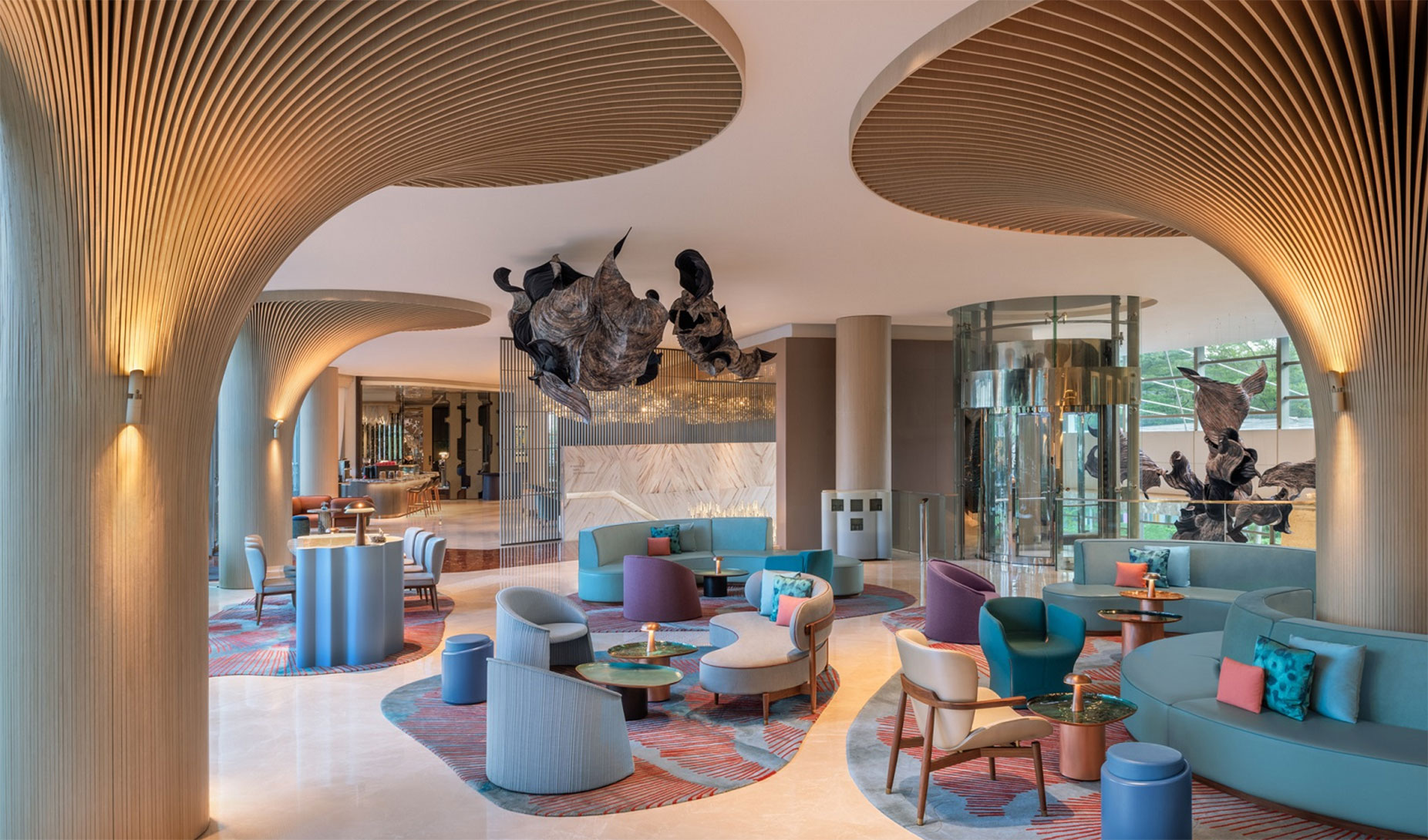 Eine moderne Hotellobby mit geschwungenen Holzsäulen, stilvollen blauen und pastellfarbenen Sitzgelegenheiten, runden Tischen, farbenfrohen Teppichen, abstrakten Deckenskulpturen und großen Fenstern, die natürliches Licht hereinlassen.