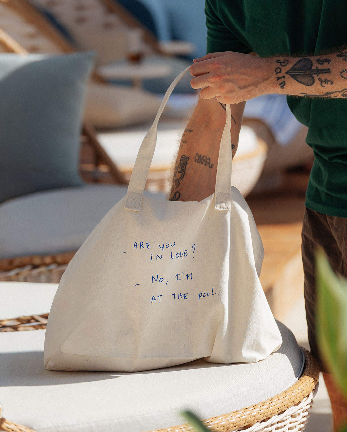 Eine Person mit tätowierten Armen stellt eine cremefarbene Tragetasche auf einen gepolsterten Stuhl. Auf der Tasche steht in blauer Handschrift: 