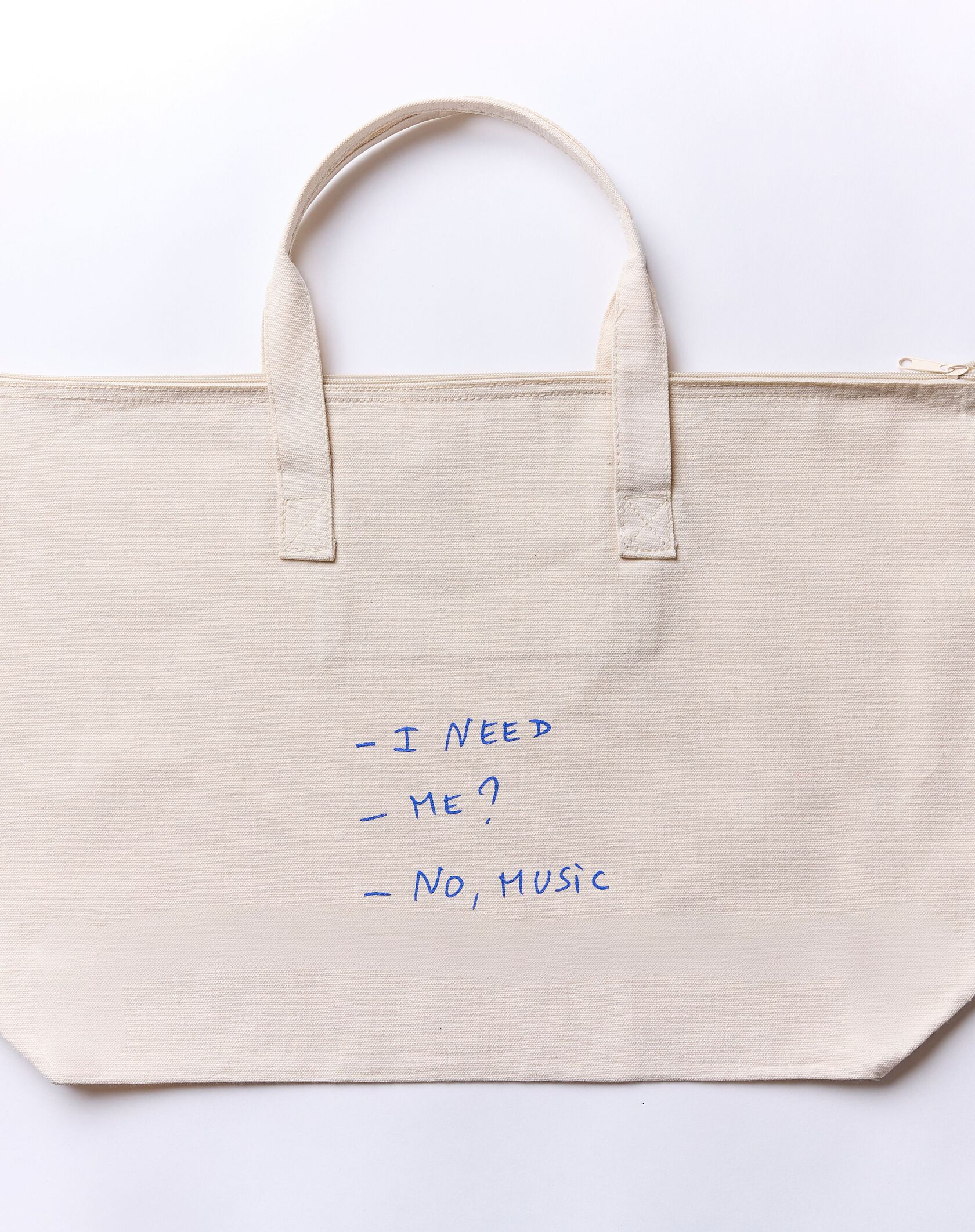 Eine beigefarbene Tragetasche aus Segeltuch mit blauem handgeschriebenem Text, der lautet: - I NEED - ICH? - NEIN, MUSIK.