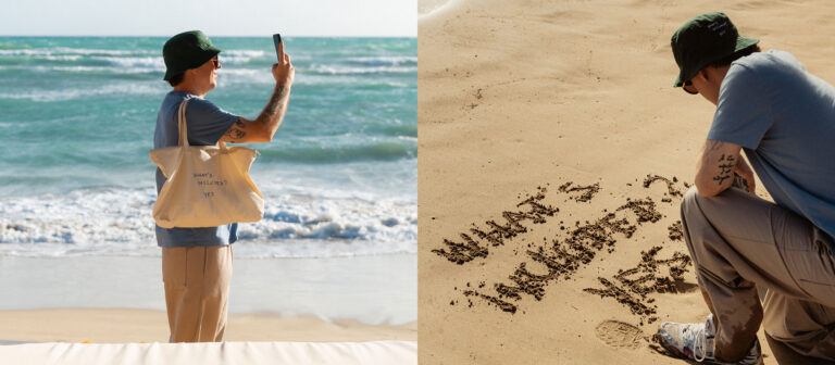Una persona en la playa: a la izquierda, de pie y haciendo una foto con un teléfono, llevando una bolsa de mano; a la derecha, agachada junto a unas palabras escritas en la arena que dicen: 