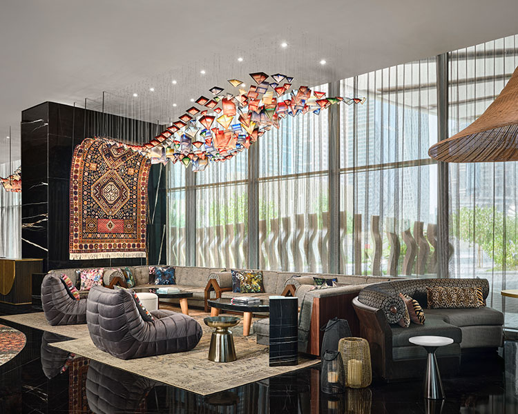Moderne Hotellobby mit stilvollen grauen Sofas, gemustertem Teppich, bunten Kissen, einzigartiger Wandkunst mit hängendem Teppich und Deckendekoration aus bunten Glasformen. Große Fenster und modernes Mobiliar schaffen einen hellen, eleganten Raum.
