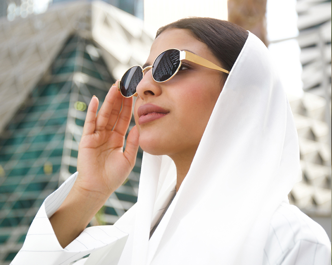A woman wearing a white headscarf and round sunglasses stands outdoors near modern skyscrapers, looking confident and stylish with her hand adjusting her sunglasses.