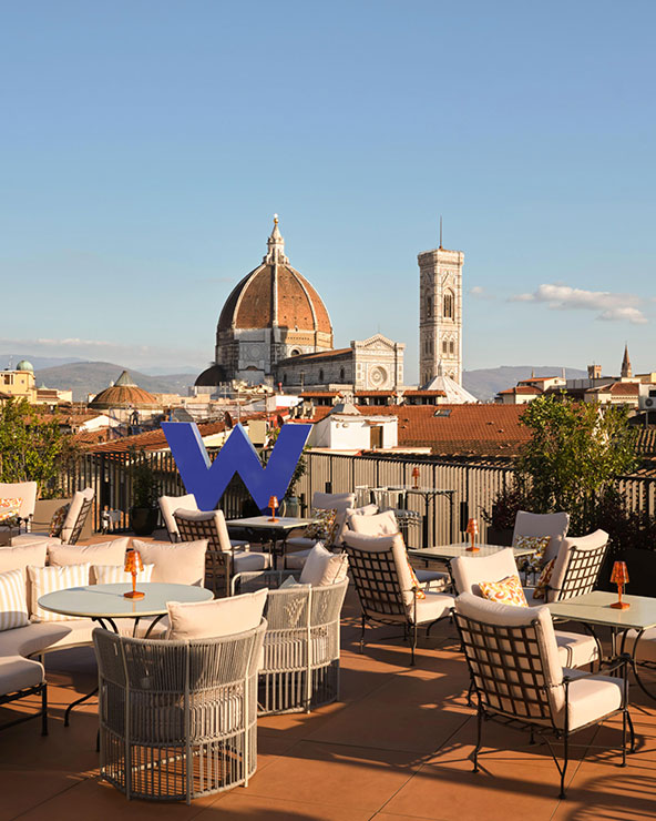 Eine Dachterrasse mit Liegestühlen und Tischen bietet bei klarem Himmel einen Blick auf den Dom und den Glockenturm von Florenz. Ein großes blaues 