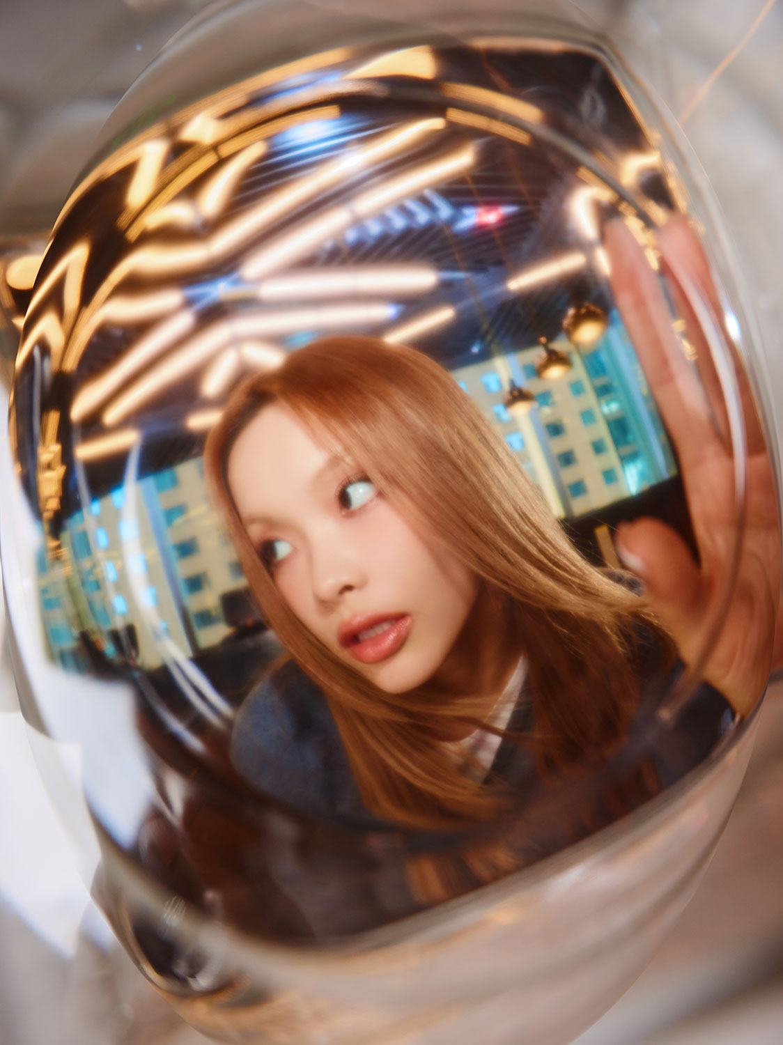 A young woman with long hair is seen through a glass sphere, creating a distorted effect. She looks to the side with her hand raised, against a background of bright ceiling lights and city windows.