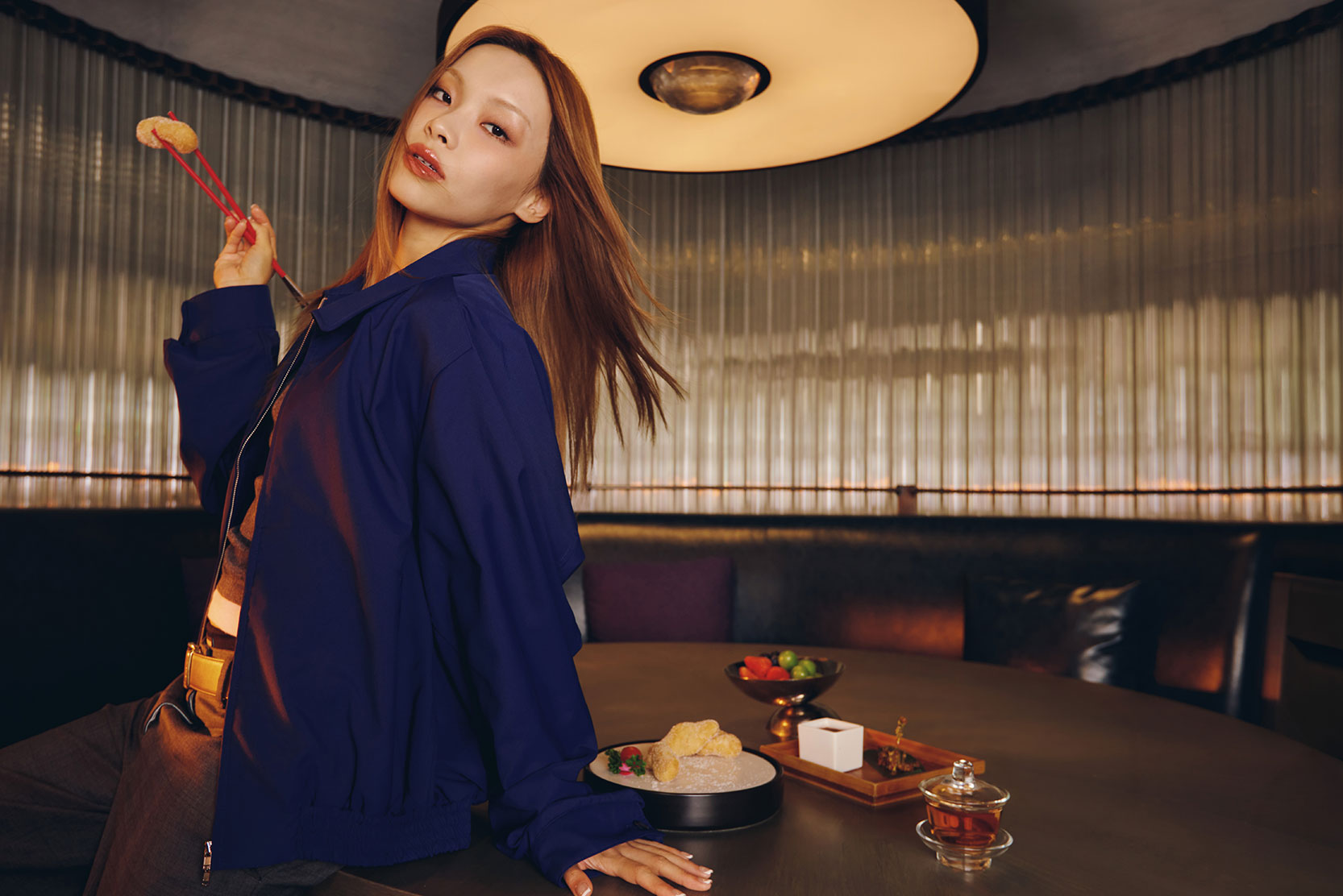 A woman in a blue jacket poses with chopsticks holding food, sitting on the edge of a round dining table set with snacks and tea, in a modern, dimly lit restaurant.
