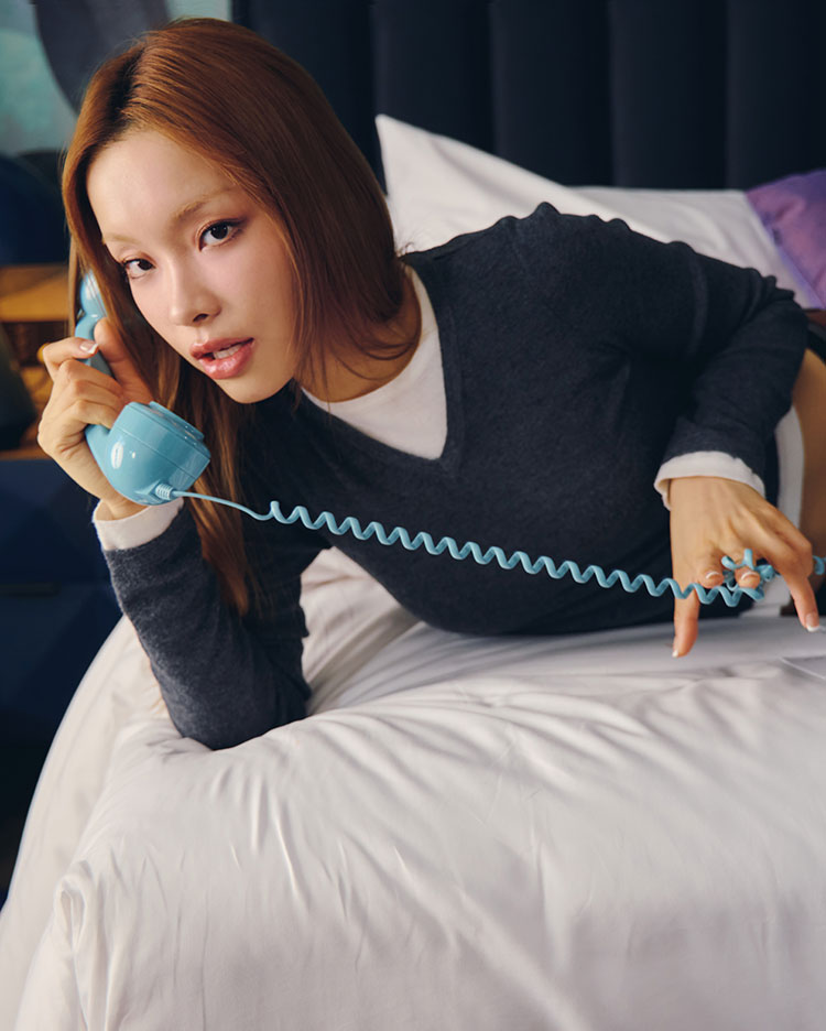 A woman with long hair leans on a bed while holding a blue corded telephone to her ear, looking toward the camera. She wears a dark sweater over a white shirt.