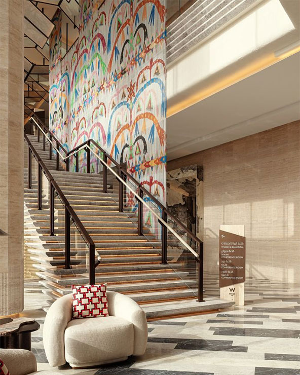 Modern hotel lobby with a wide staircase, colorful abstract wall art, a rounded beige chair with a red-and-white cushion, and sunlight streaming through large windows.