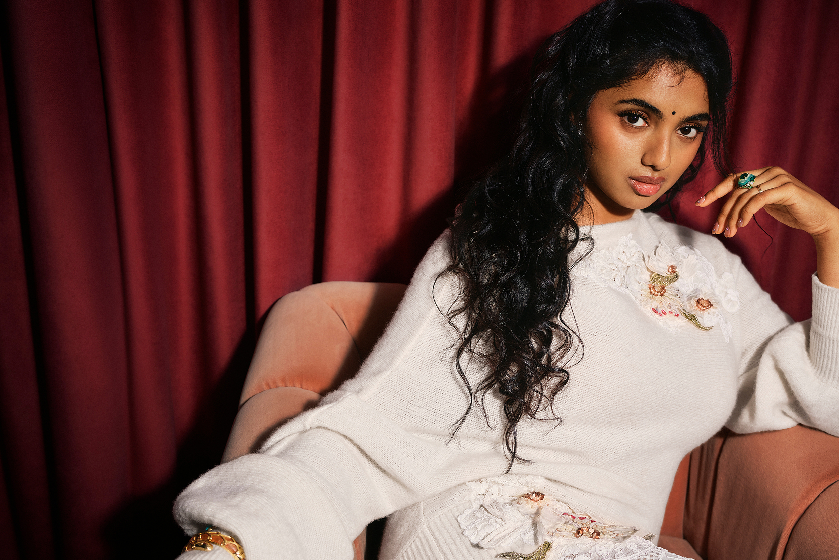 A woman with long dark hair sits in a pink chair, wearing a white sweater with floral embroidery, against a backdrop of dark red curtains.